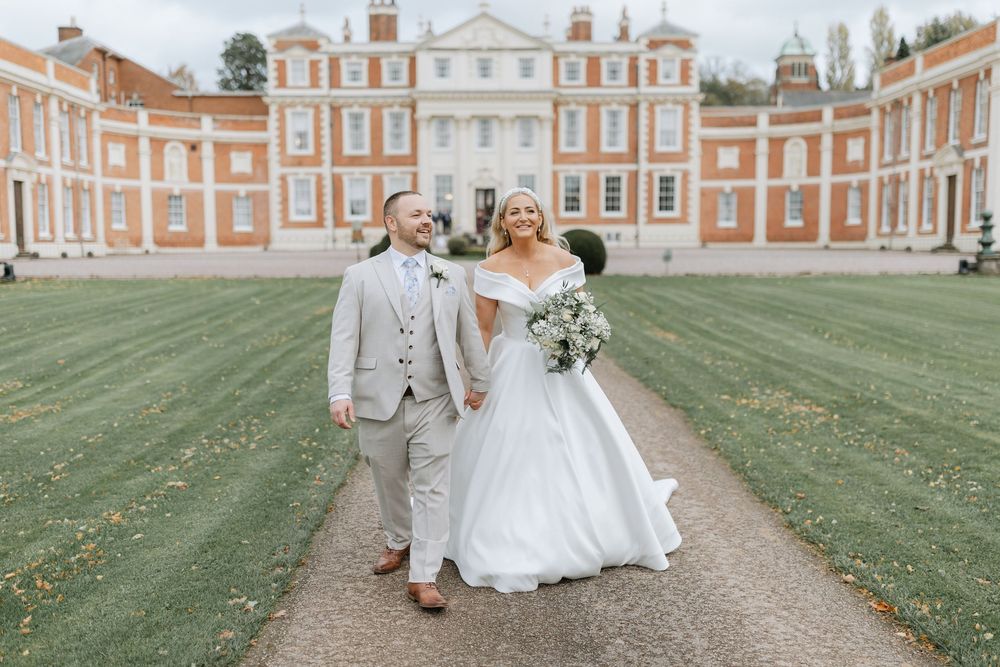 Couple in wedding attire walking together on a path through a grand English estate garden with historic brick buildings.