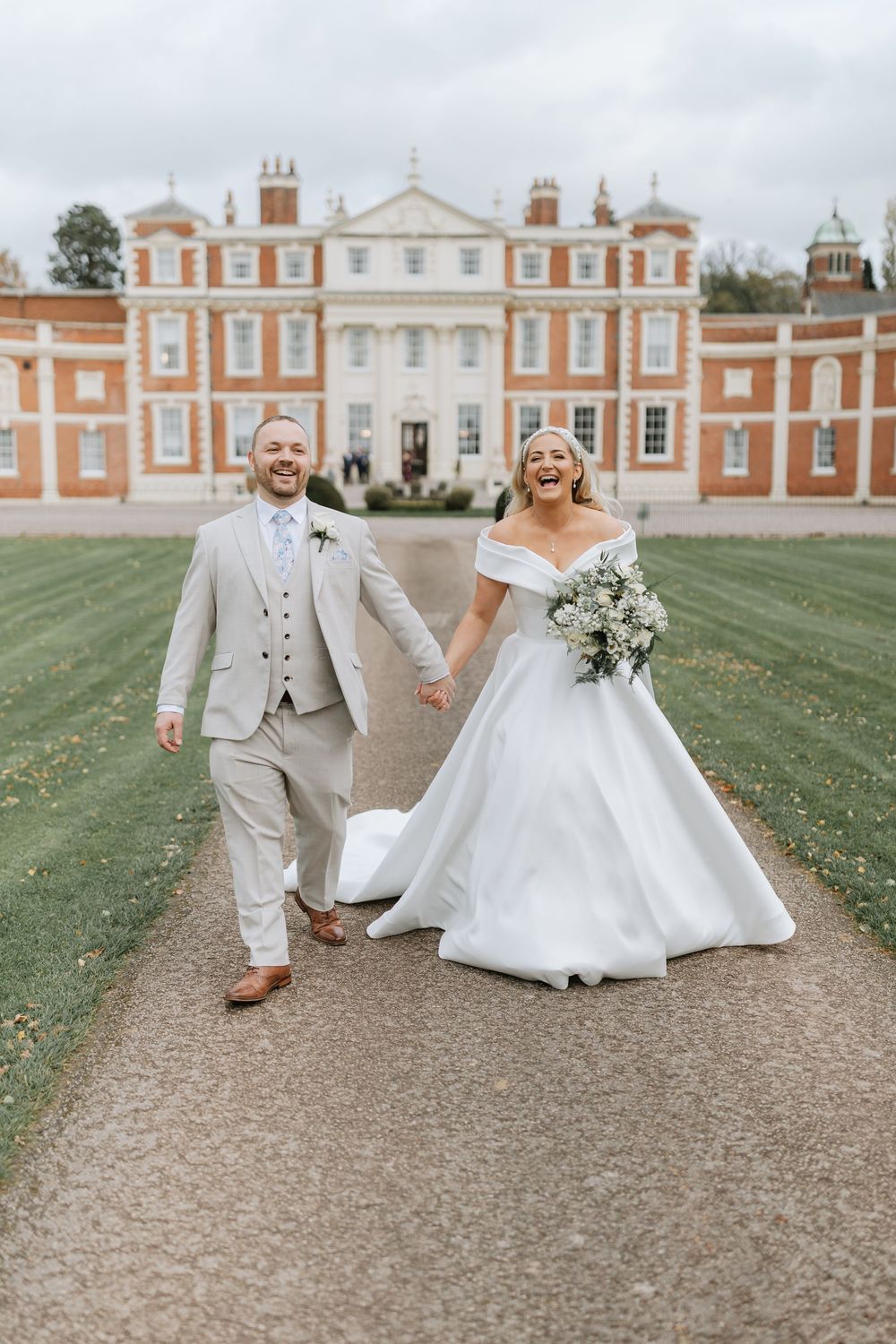 A couple in wedding attire joyfully walks along a gravel path in front of a grand historic mansion.