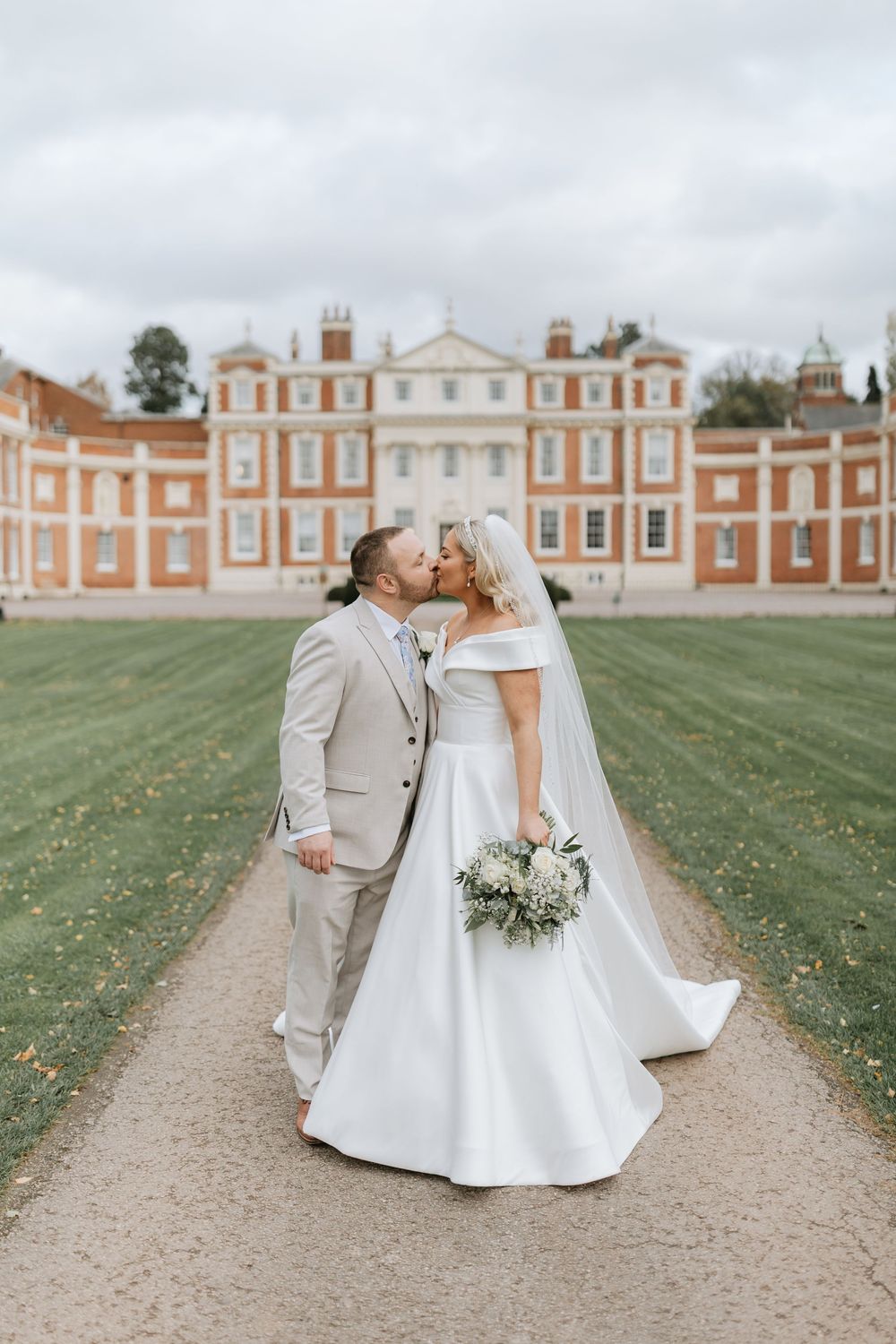 Wedding couple poses on garden path with historic red brick estate building creating a romantic backdrop.
