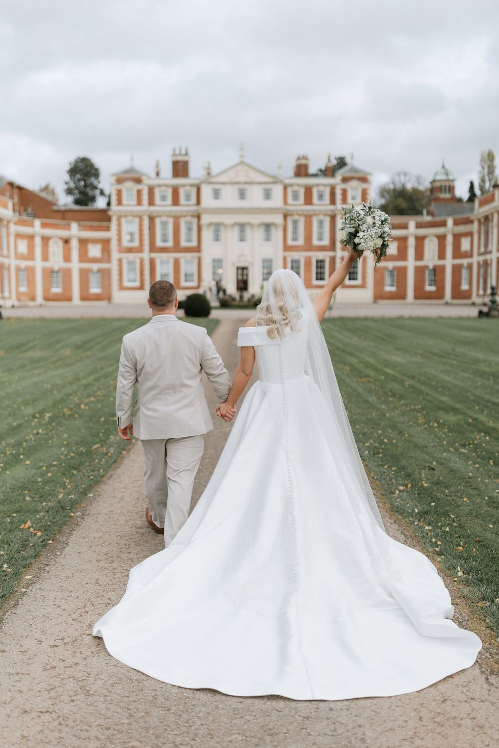 Couple in wedding attire walking along a gravel path with elegant brick mansion and manicured grounds in background.
