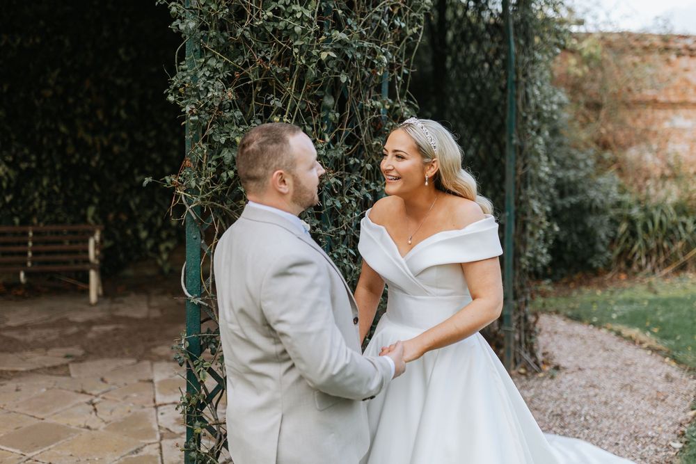 A wedding couple walks hand in hand through a rustic garden pathway surrounded by greenery and brick walls.