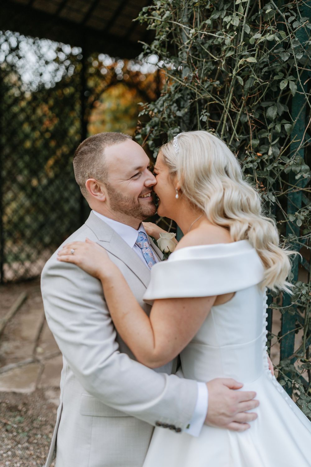 A romantic wedding moment captured in a garden setting with a couple in white formal attire sharing an intimate embrace.