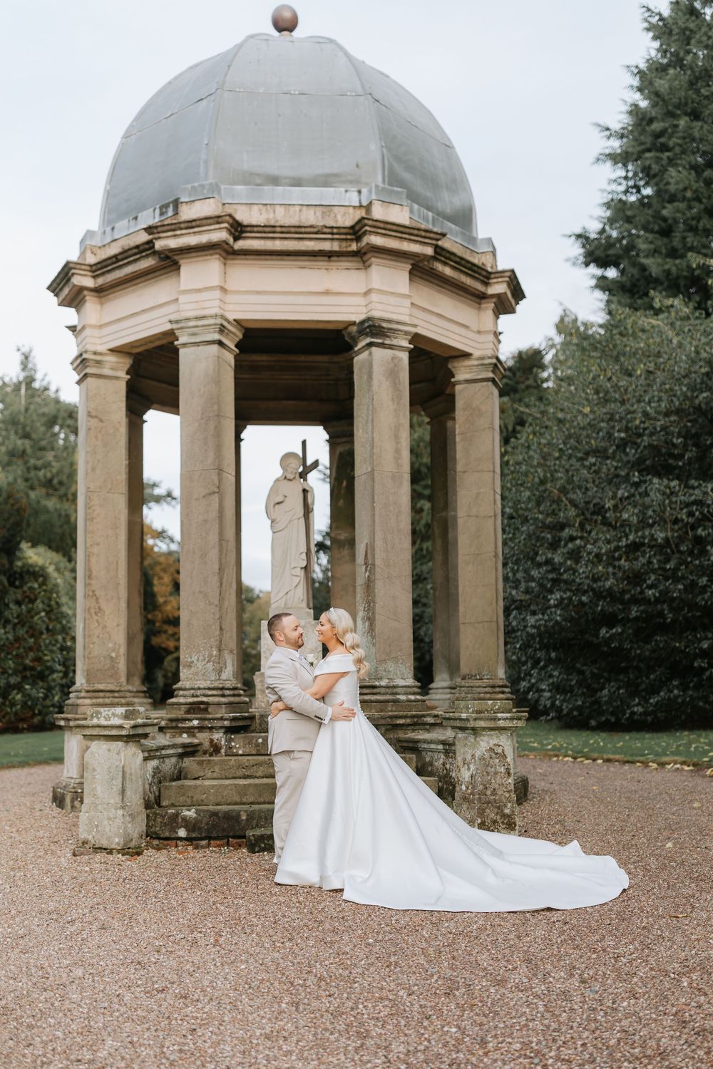 Elegant stone gazebo with domed roof surrounded by trees on gravel path at historic estate garden.