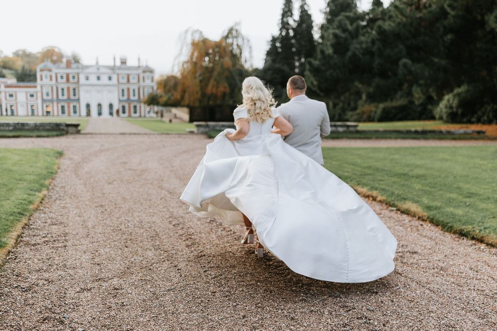 A couple in wedding attire walks along a gravel path surrounded by manicured lawns and a grand estate house.