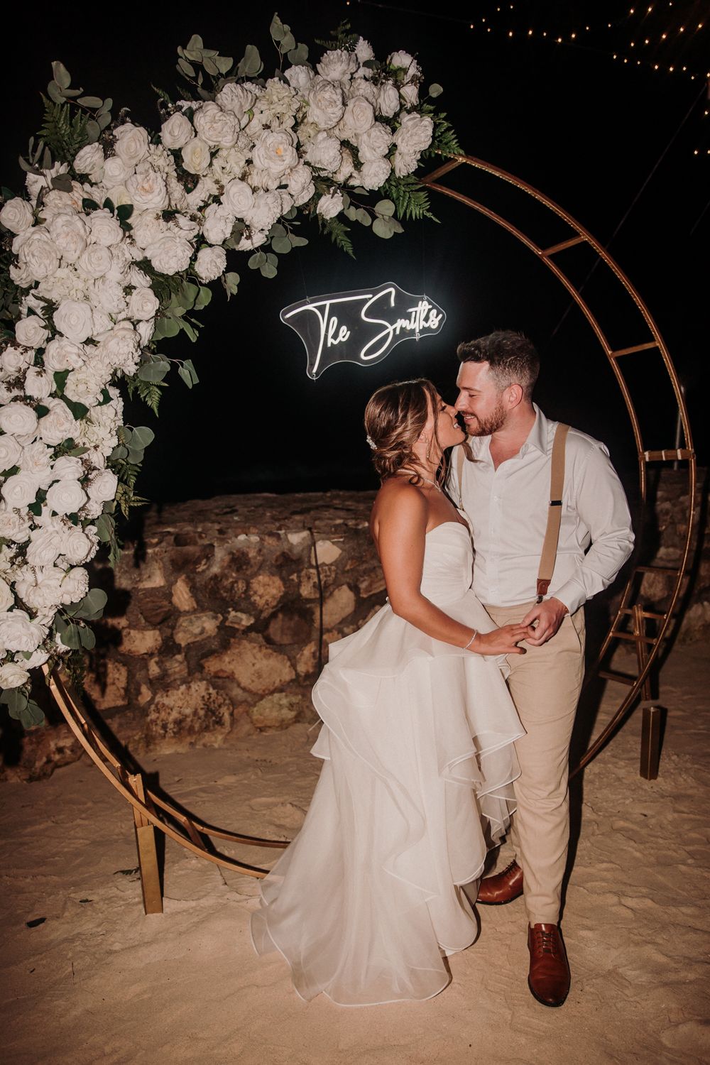 A couple poses in front of a floral arch with white orchids on a sandy beach at night. El Dorado Royale Wedding Photographer