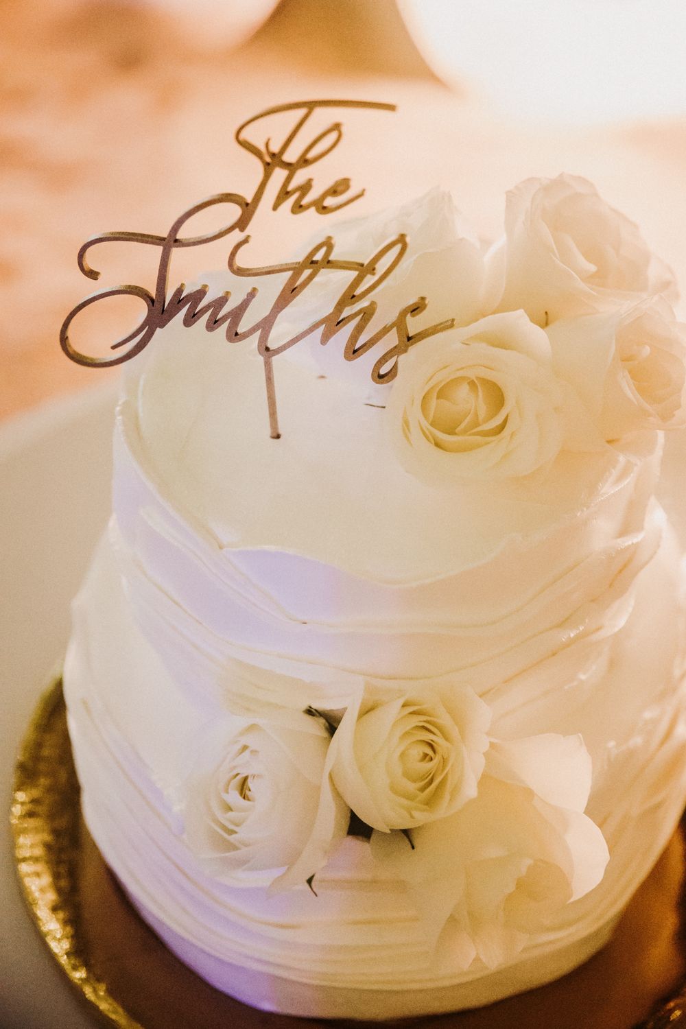 Elegant white wedding cake decorated with cream roses and cursive "The Smiths" topper on gold base. El Dorado Royale Wedding Photographer