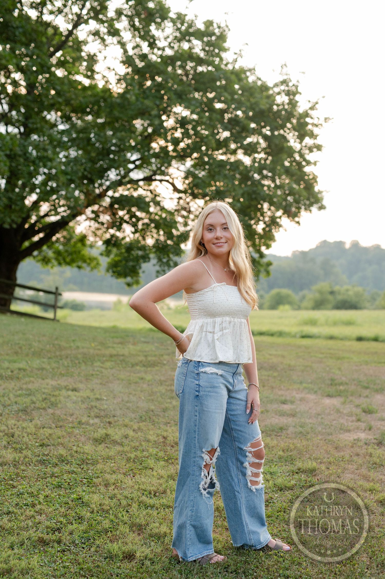 Summer fashion portrait against pastoral landscape with fence and trees.