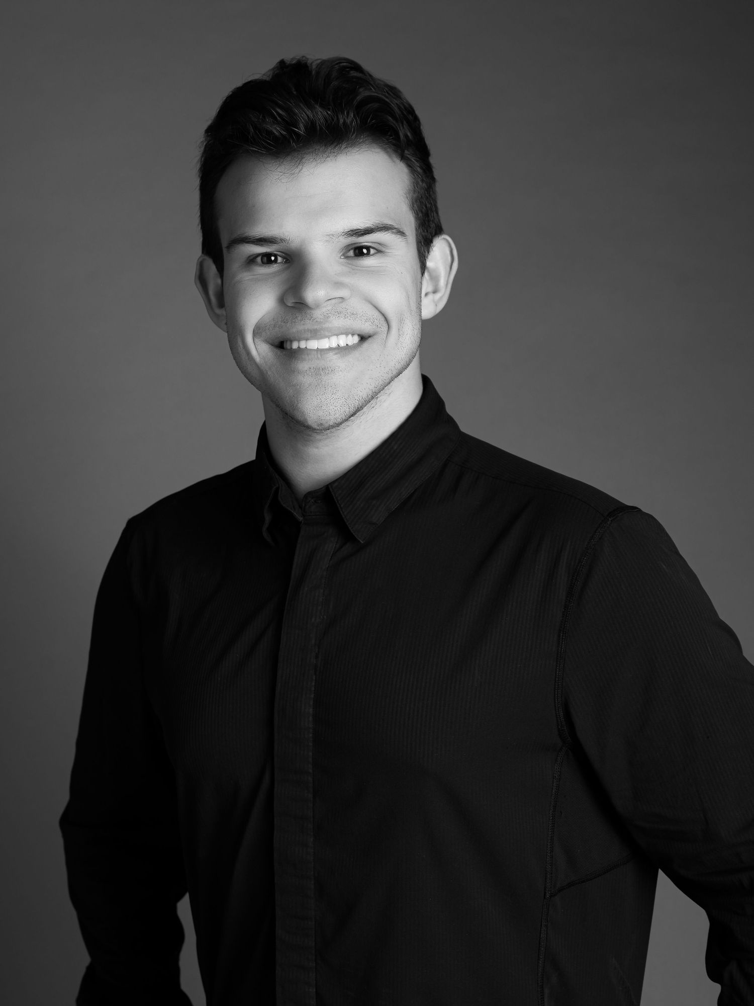 Professional black and white headshot of a person in a dark button-up shirt smiling warmly against a neutral backdrop.