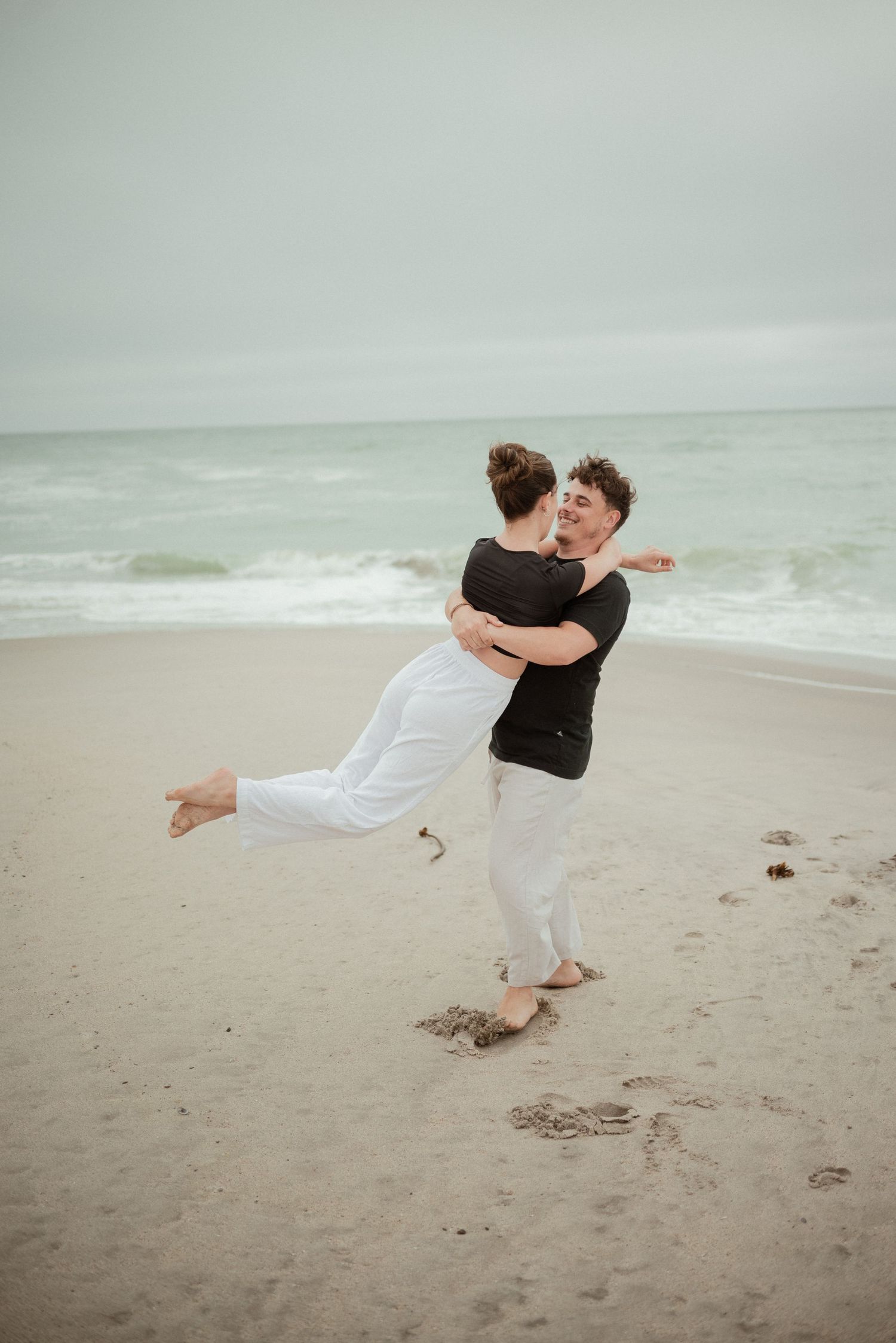 A couple embraces joyfully on a cloudy beach with waves rolling in the background.