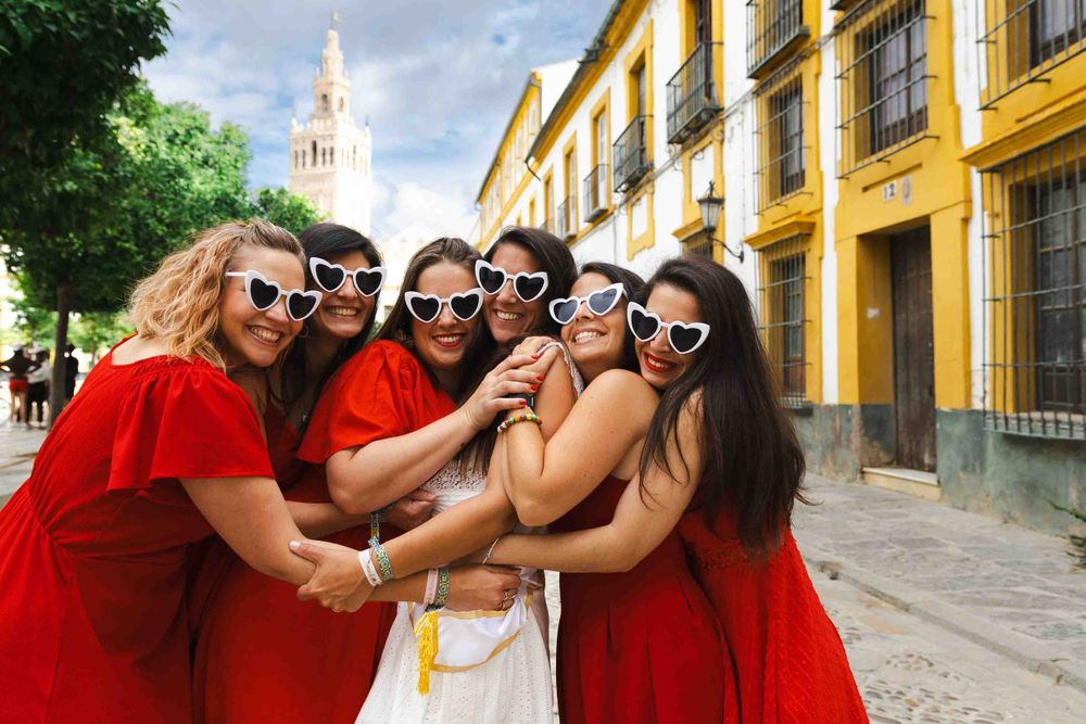 A group in matching red dresses and sunglasses gather around a person in white on a sunny Spanish street.