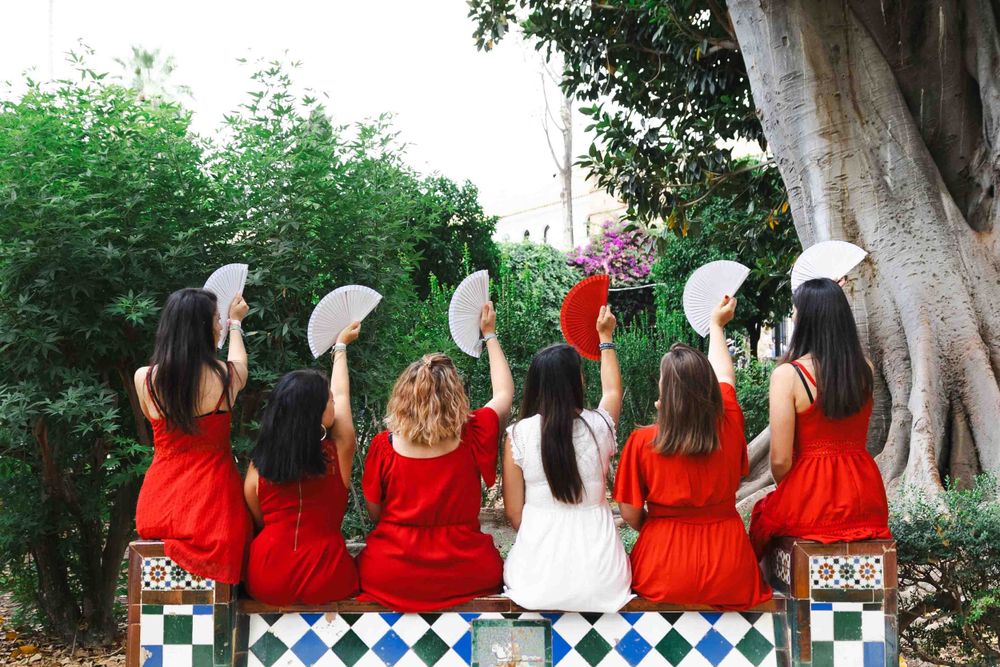 A group in matching red dresses sit together on a tiled bench by a large tree trunk in a garden setting.