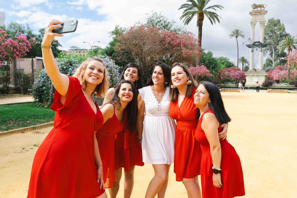 A group in coordinating red dresses and one in white pose for a selfie in a sunny garden with palm trees.