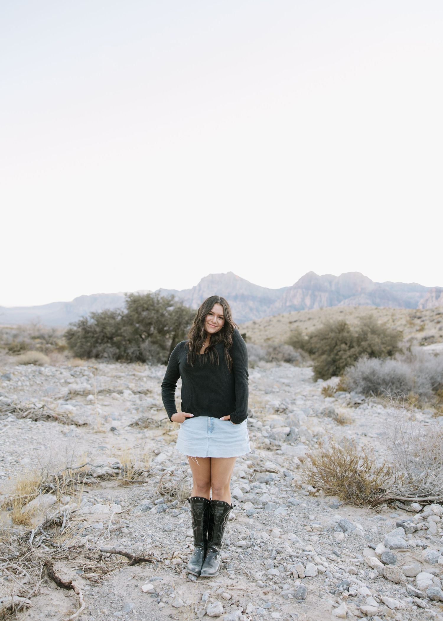 Woman in black sweater and white skirt poses in desert landscape with mountains and brush in background.