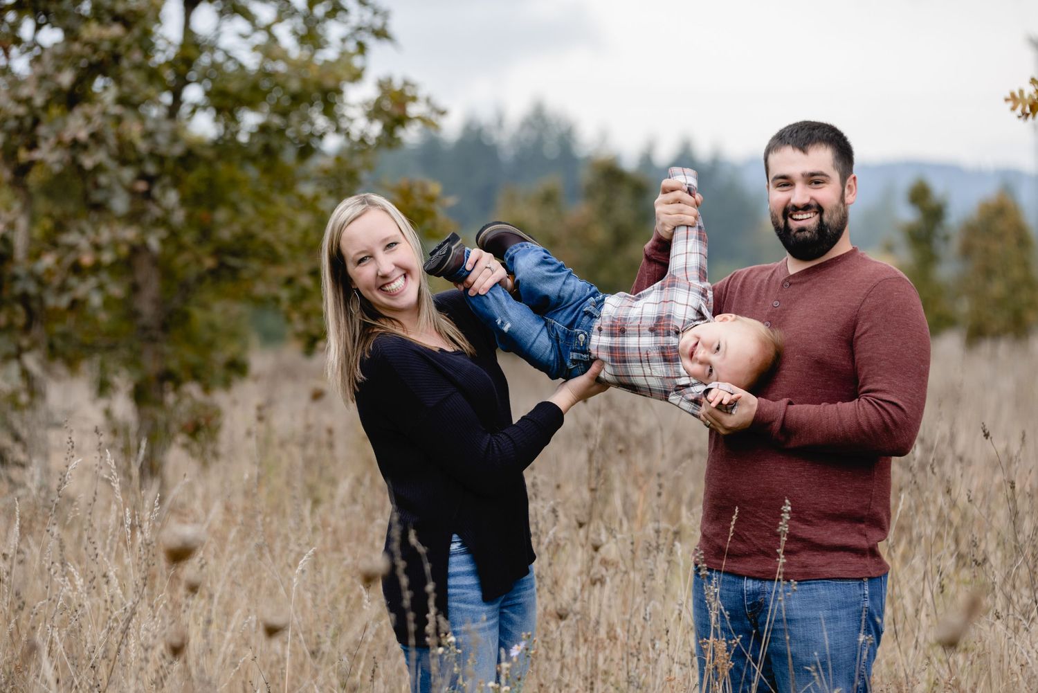 A playful family moment captured in a grassy autumn field with fall foliage in the background.