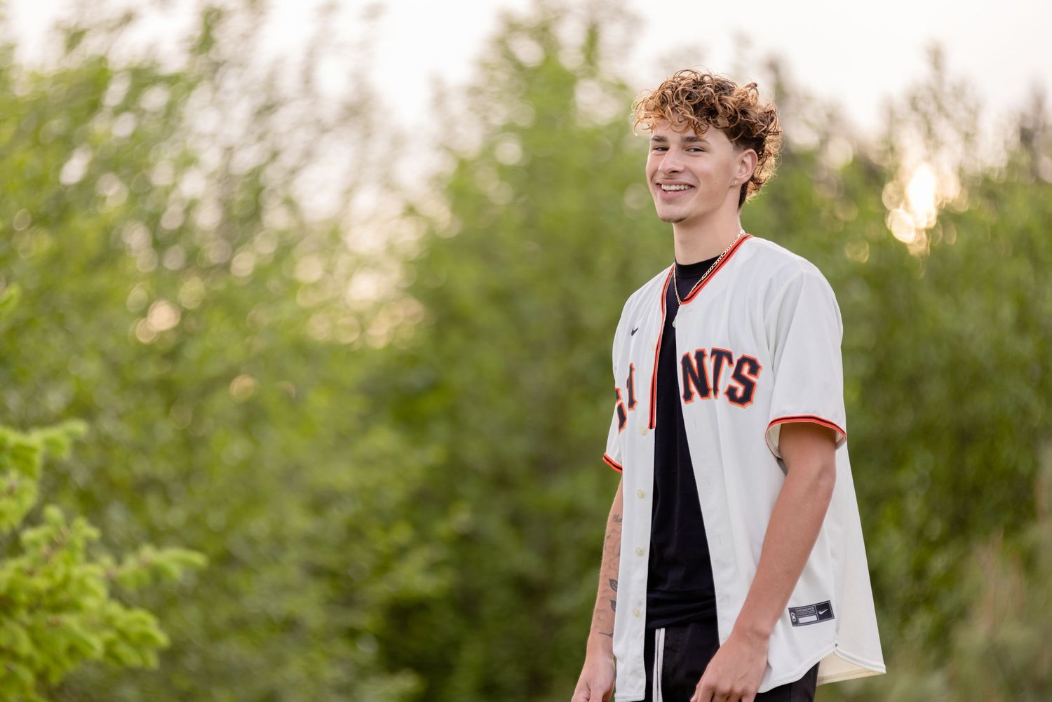 A young person wearing a white Giants baseball jersey stands outdoors in natural lighting during golden hour.