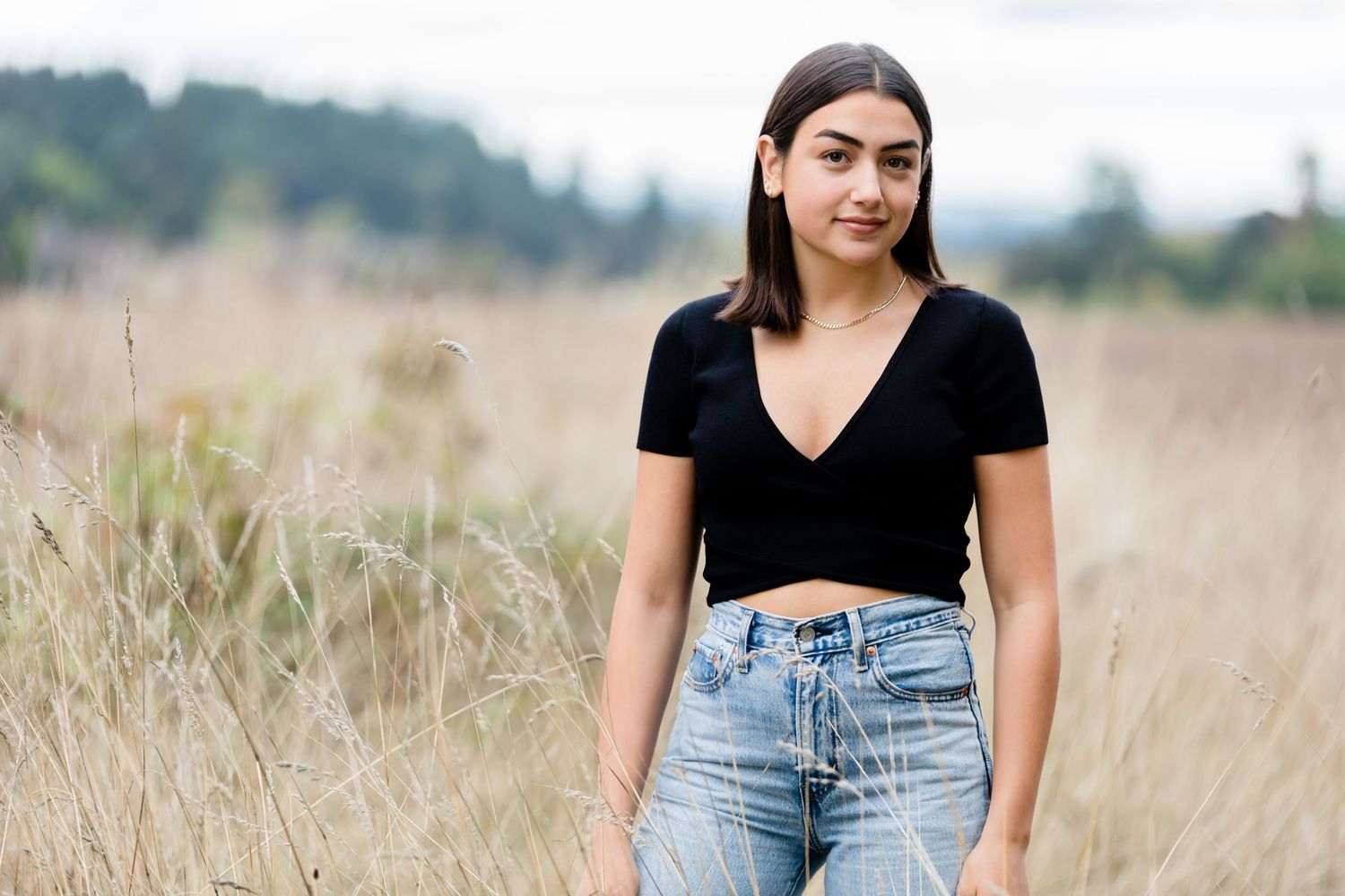 Someone wearing a black crop top and high-waisted jeans stands in a field of tall grass on a summer day.