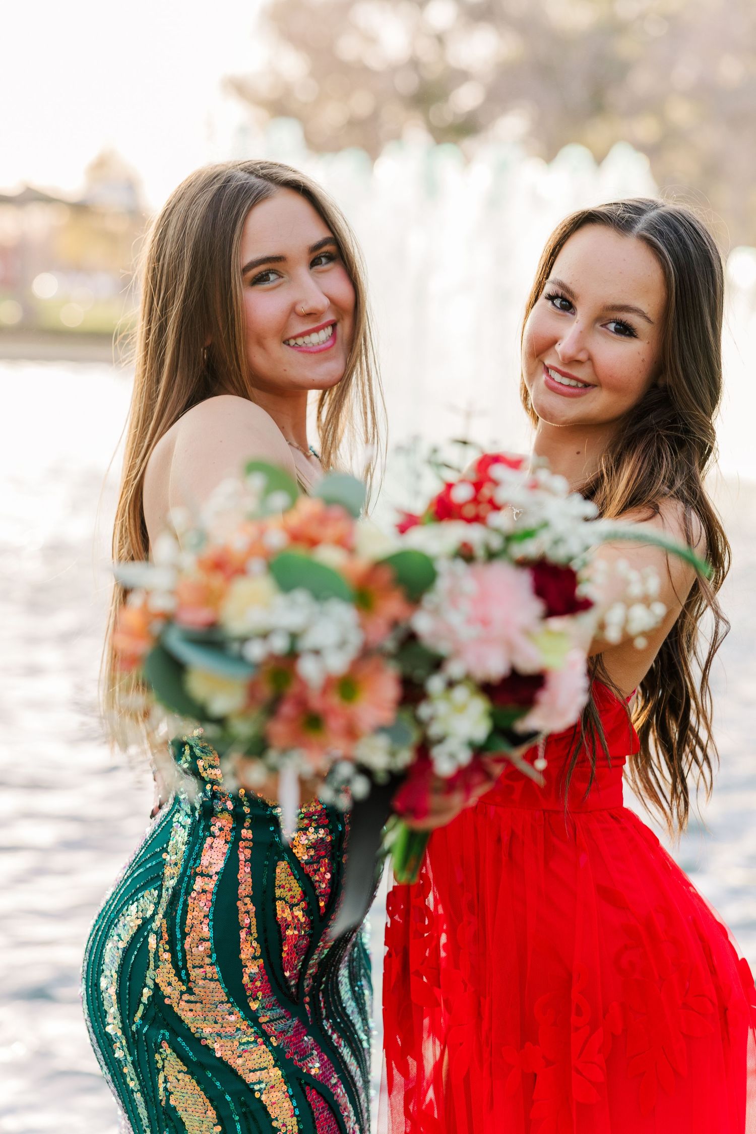 Two friends in formal dresses, one in green sequins and one in red, holding colorful floral bouquets outdoors.