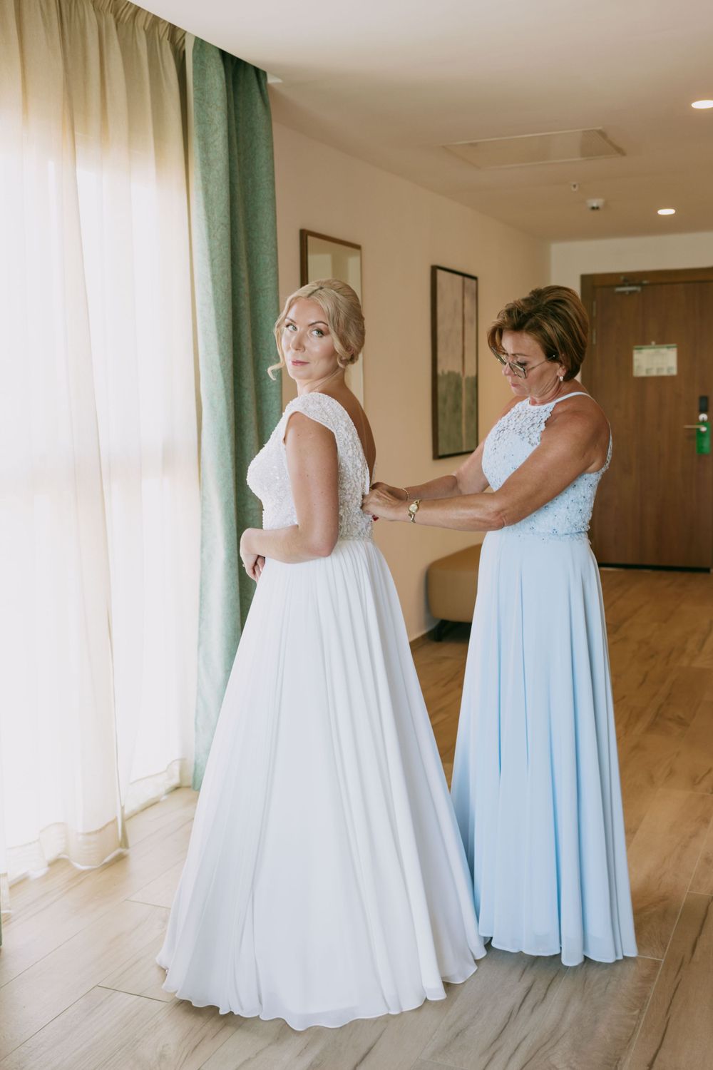 Two people share an intimate moment during wedding dress preparations in a sunlit room.