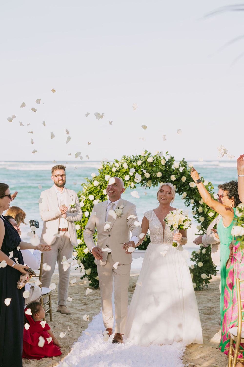 Joyful beach wedding ceremony with guests throwing petals under a floral arch.