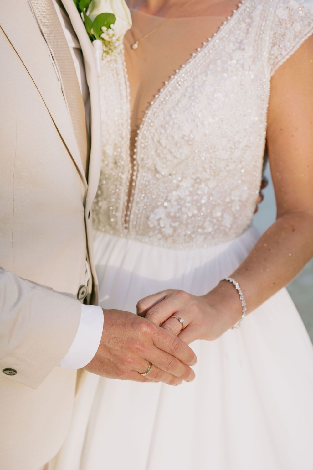 Close-up of wedding couple's hands showing rings and detailed white dress.