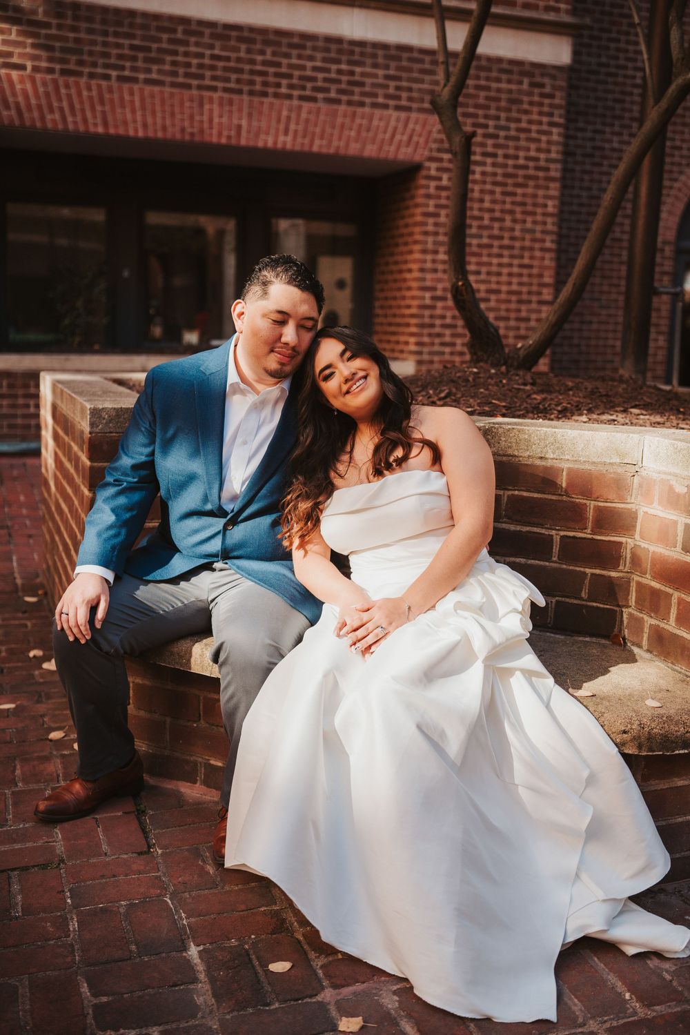 A bride in a white wedding dress and groom in a blue suit sit together on brick steps outside a building.