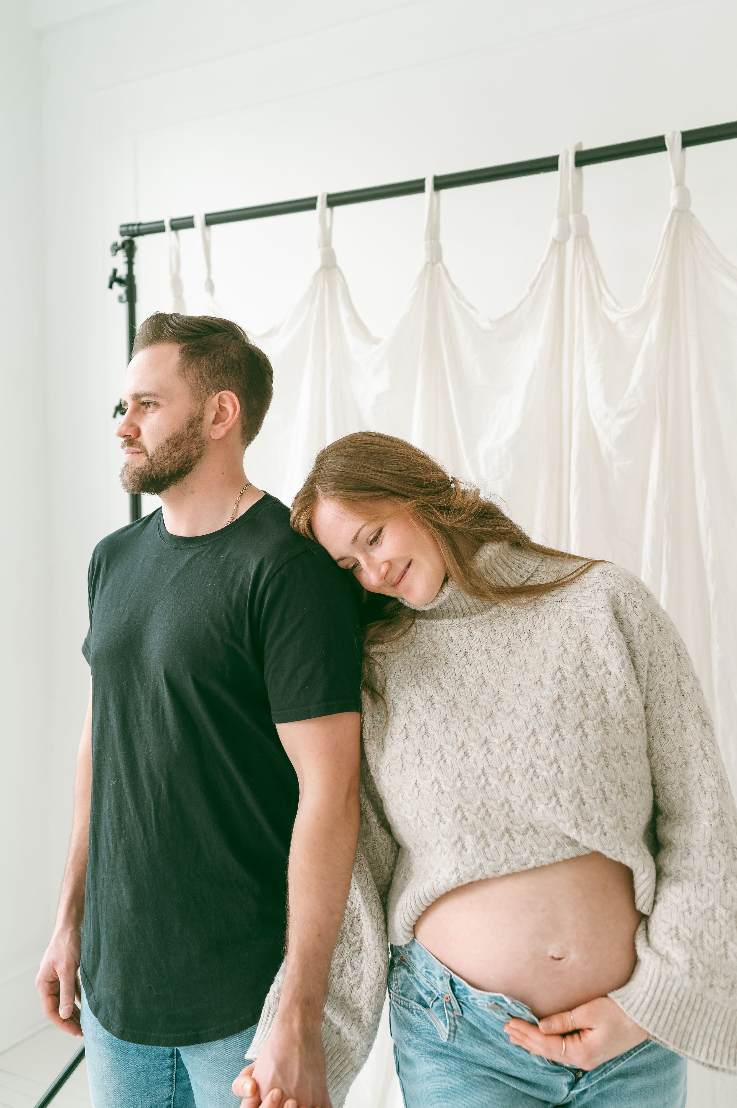 A couple stands together against a white backdrop while the woman shows her pregnant belly in a cozy knit sweater.
