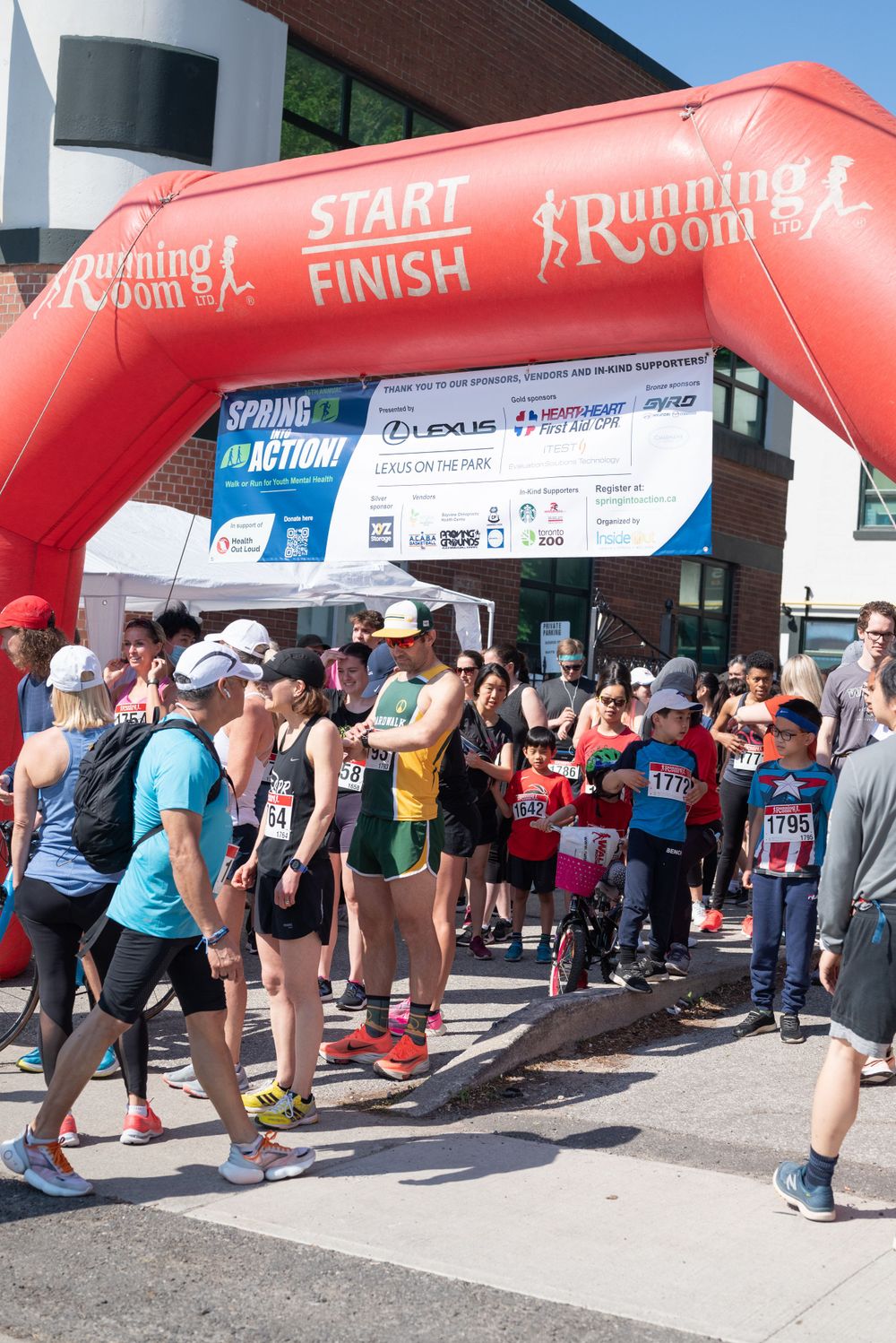 Runners gather at red inflatable start/finish line arch at a Running Room race event on a sunny day.