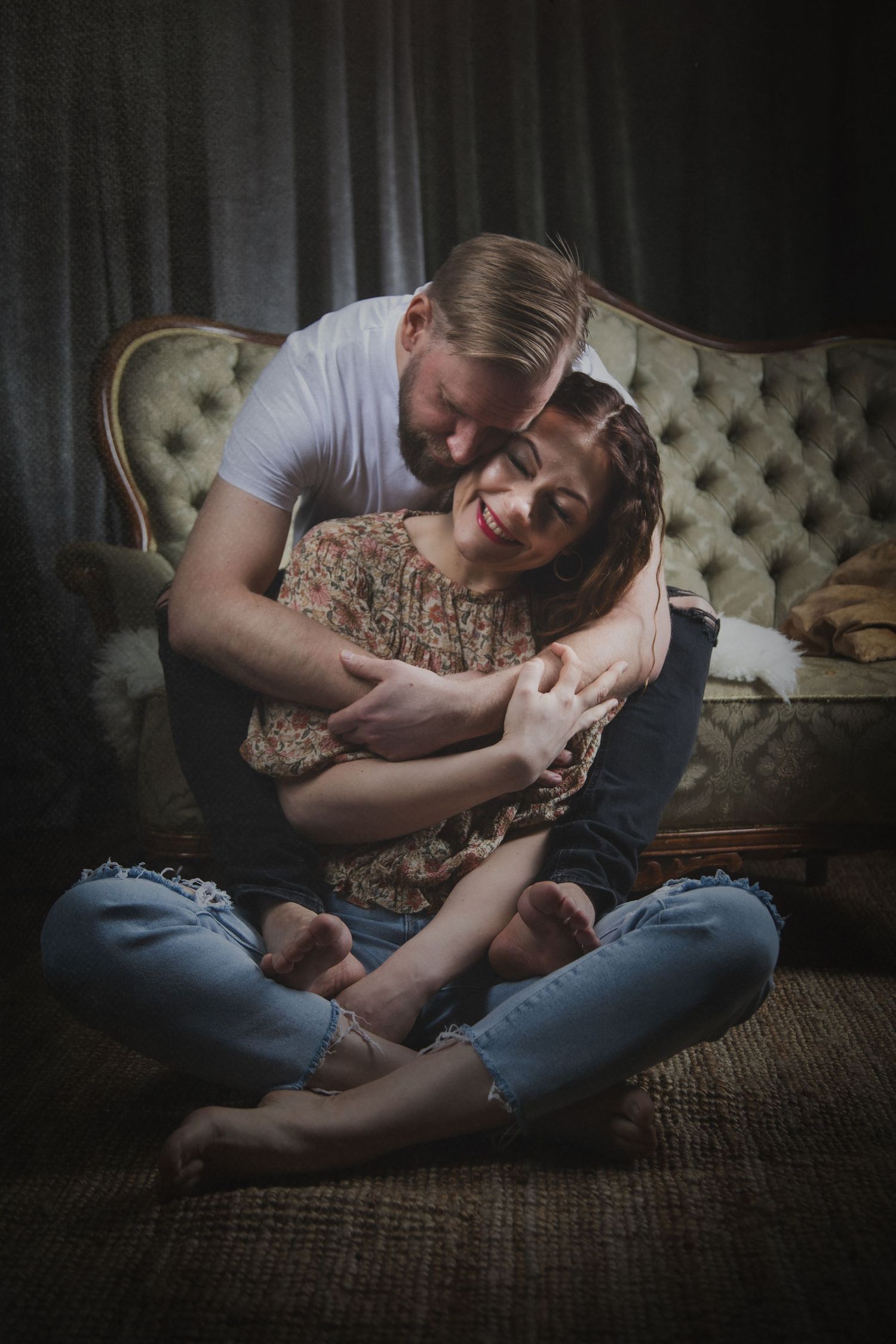 Intimate indoor portrait of couple cuddling on tufted vintage couch in moody lighting.