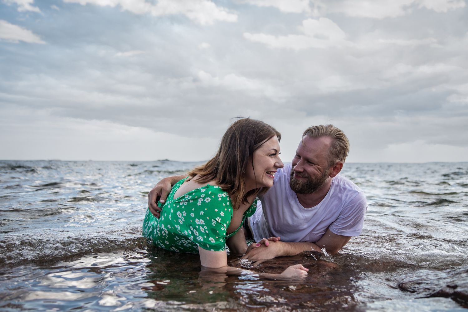 Joyful couple laughing together while playing in ocean water on cloudy day.