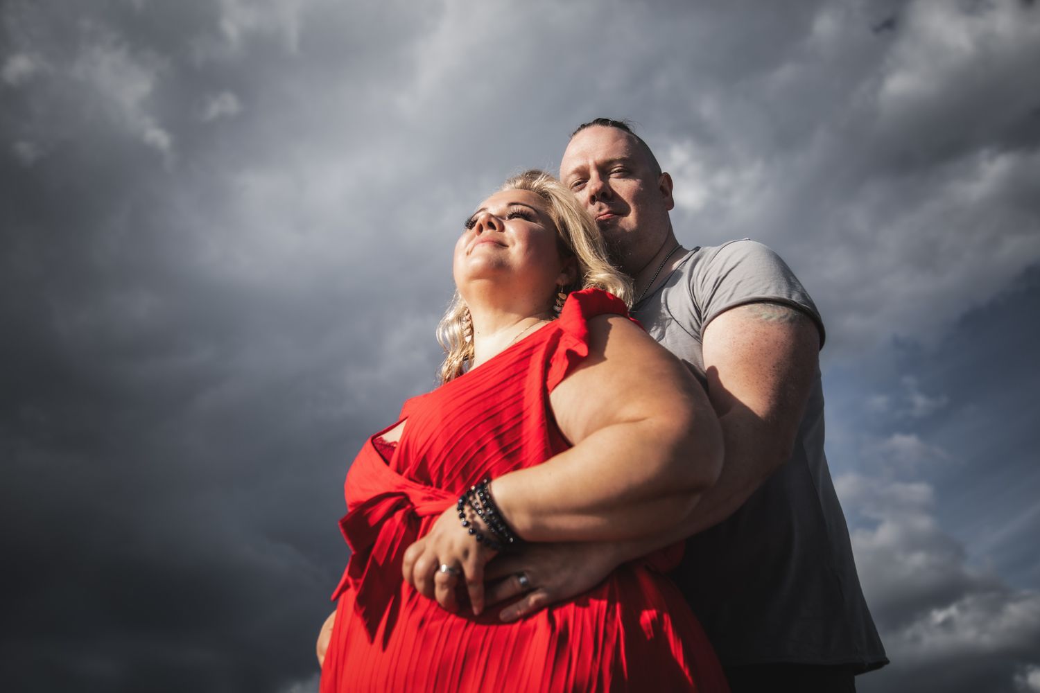 Dramatic portrait of couple embracing against stormy sky with woman in flowing red dress.