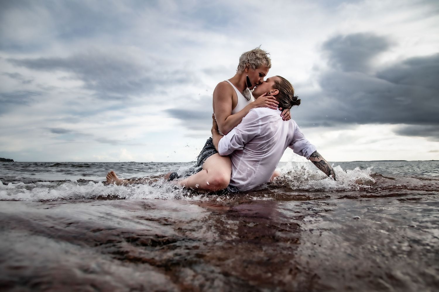 A romantic couple embraces while wading in water at the beach under dramatic cloudy skies.