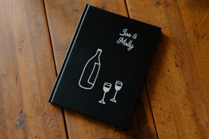 Black hardcover book with minimalist wine bottle and glasses illustration on wooden surface.