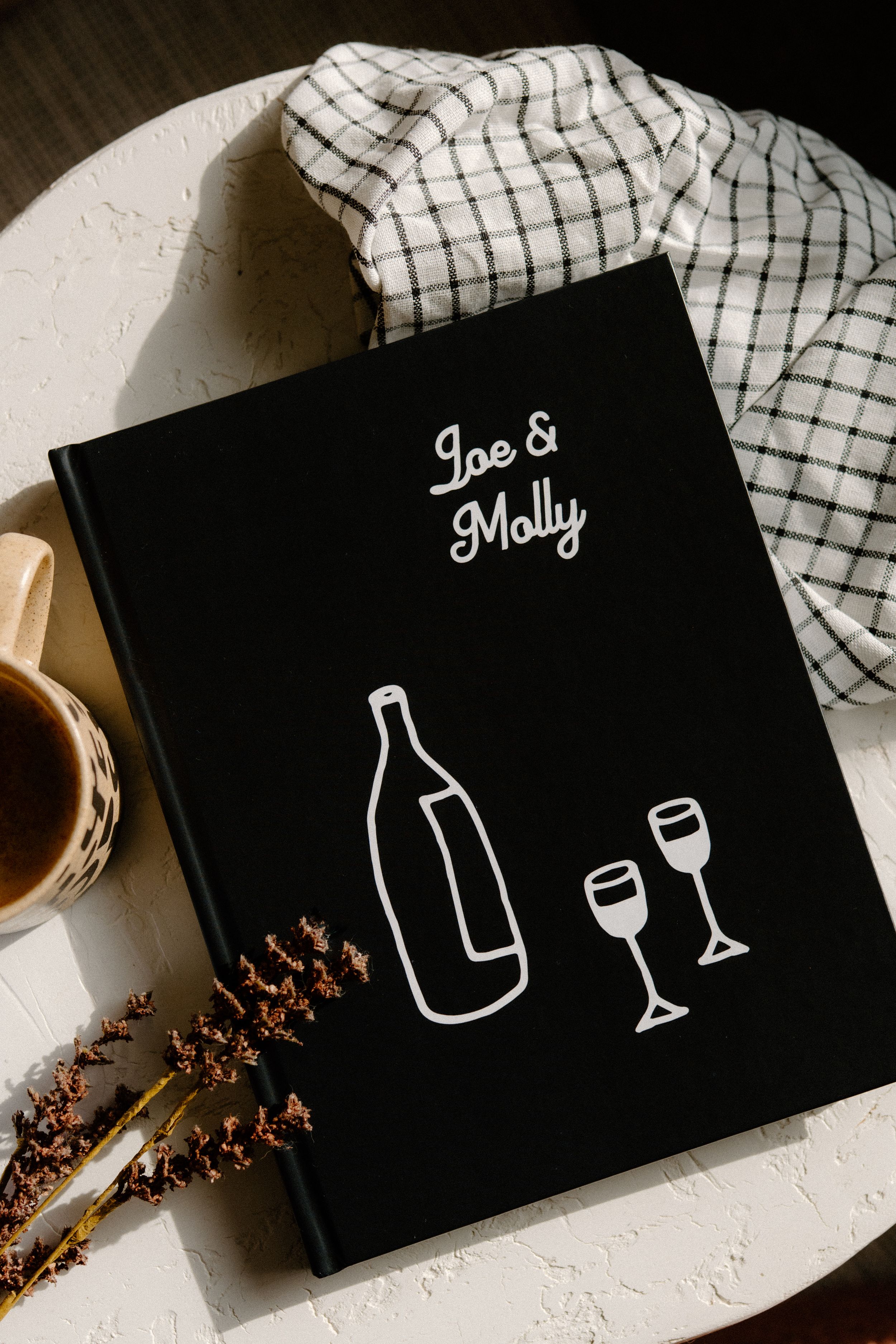 Black book cover with wine bottle and glasses illustration, names Joe & Molly on display with checkered cloth backdrop.