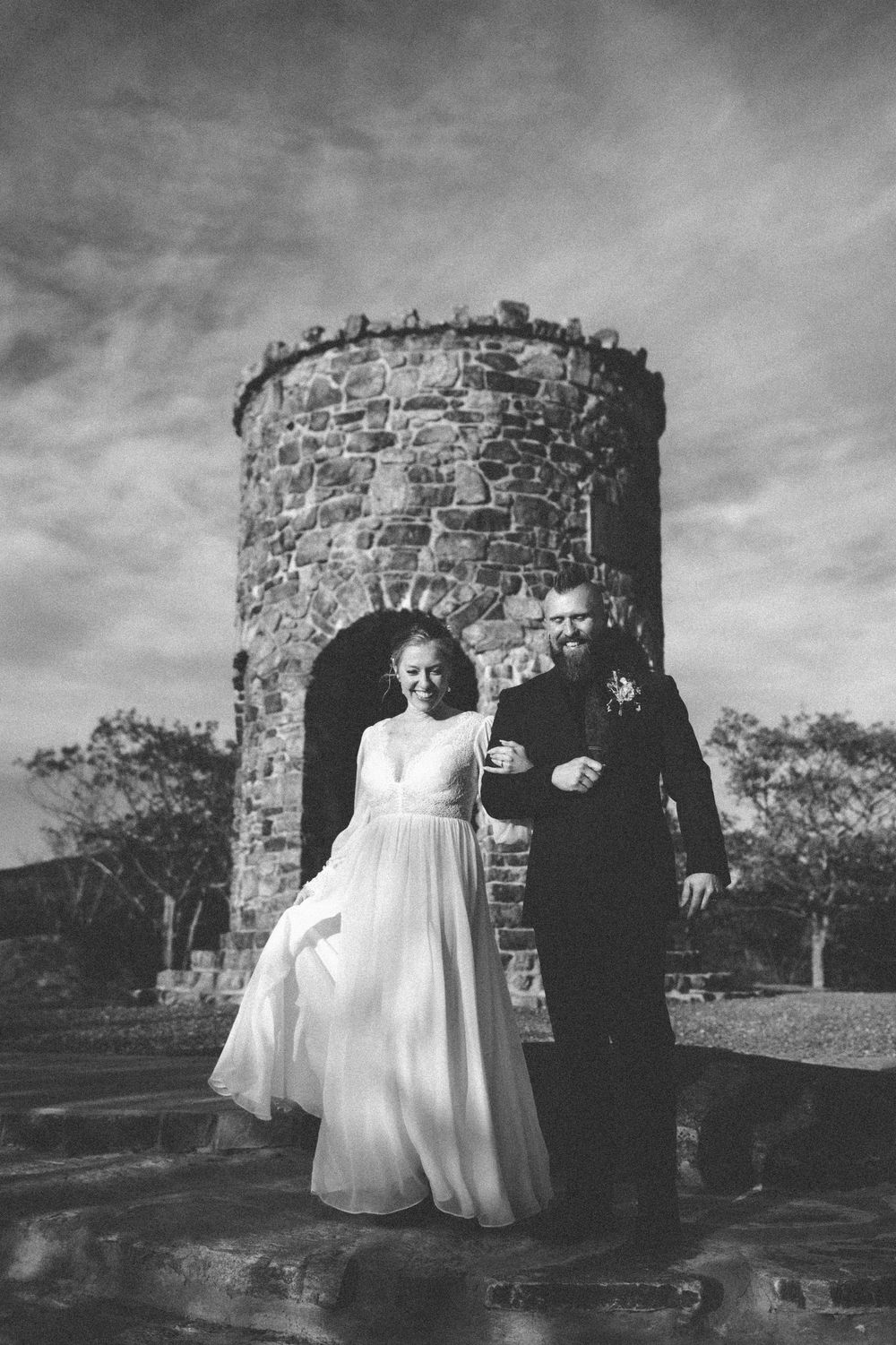 Black and white wedding photo of bride and groom walking arm in arm before a rustic stone tower under dramatic cloudy skies.