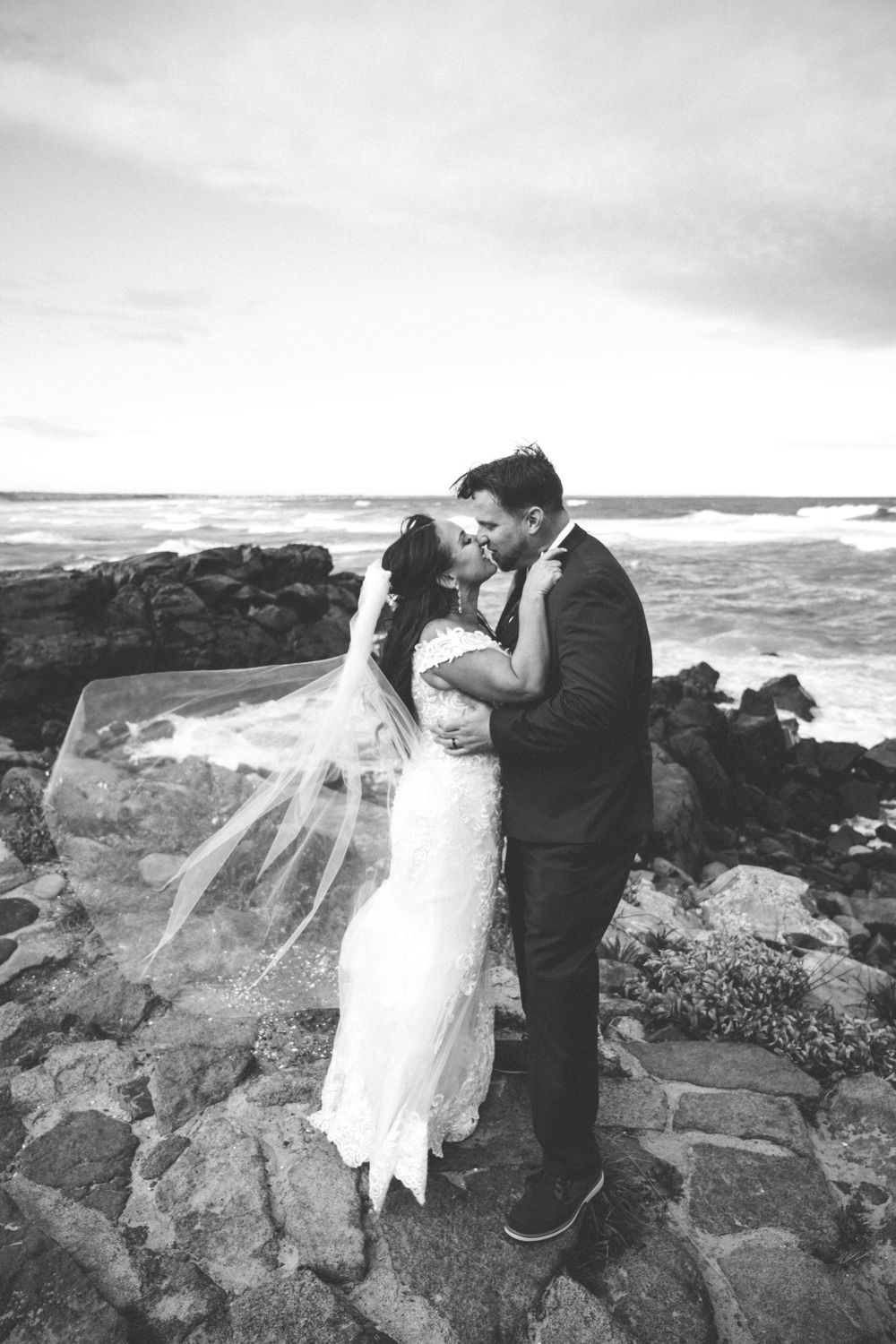 Black and white photo of a bride and groom kissing on rocky shoreline, veil blowing in wind with ocean waves behind them.