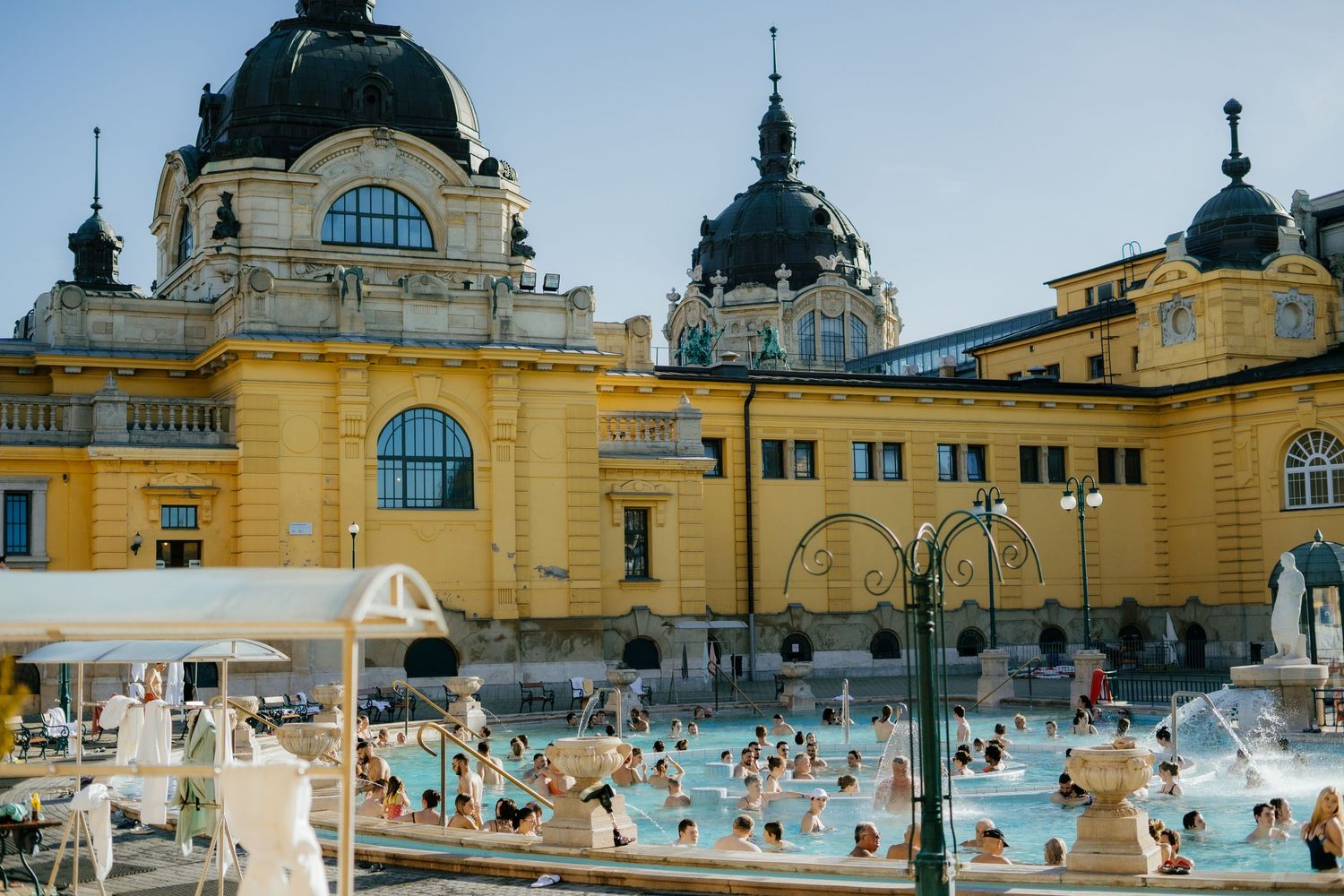 Historic yellow Szechenyi thermal bath building in Budapest with people enjoying the outdoor thermal pools.