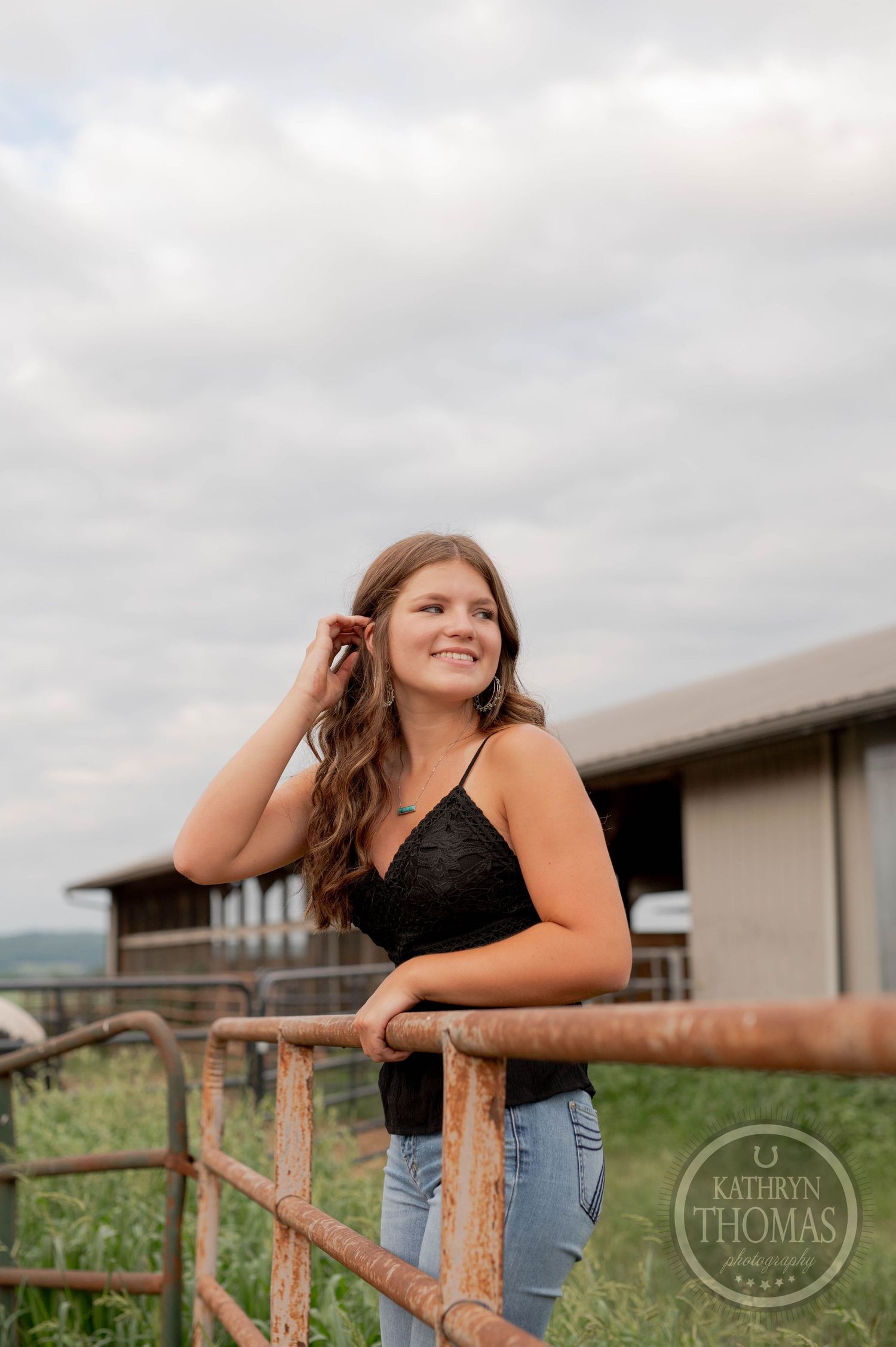 Young woman in black top leans on rustic wooden fence while posing for a casual outdoor portrait.