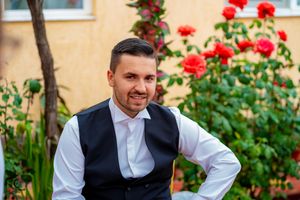 Man in formal vest and white shirt stands near blooming rose garden.