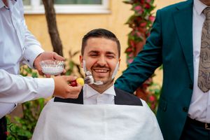 Groomsmen help adjust a bowtie during outdoor wedding preparations against yellow wall with red flowers.