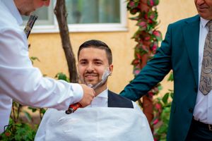 Groomsmen assist with wedding day preparations in front of yellow stucco wall with red flowers.