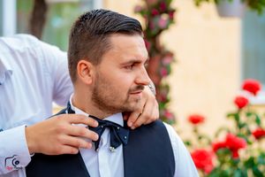 Groomsmen assist with formal wear preparations against floral decorated yellow wall.