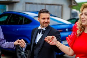 Wedding party members dance in a line outside with cars visible in the background.