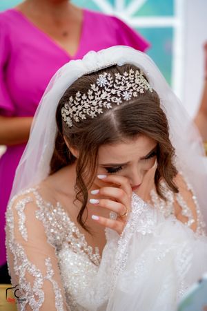 A bride wearing an ornate crystal tiara and lace dress wipes tears during an emotional wedding moment.