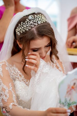 Emotional bride in crystal headpiece and lace gown reads a card while wiping away tears.