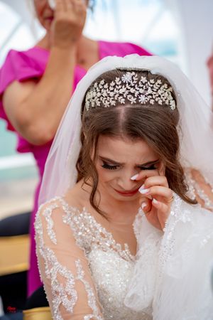 Bride wearing delicate lace dress and crystal crown wipes tears during wedding ceremony.