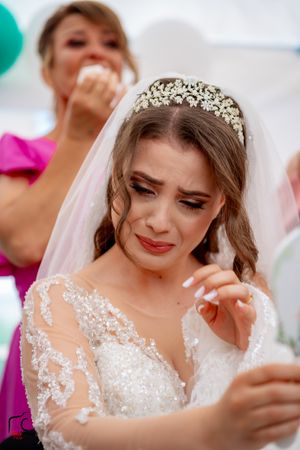 Tearful bride in elegant crystal tiara and lace gown holds something while getting emotional.