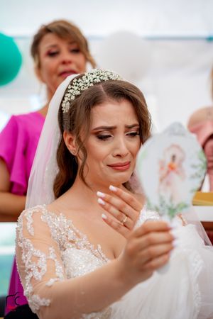 Emotional bride in crystal crown and lace dress holds card while wiping tears.