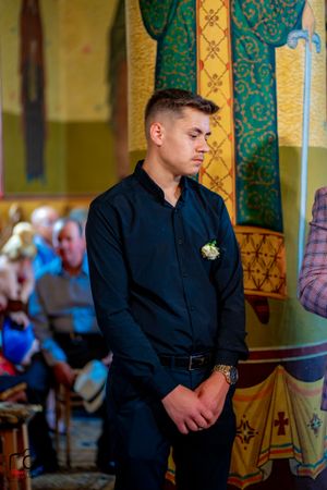 Wedding guest in a black suit stands near ornate green and gold decorative columns during ceremony.