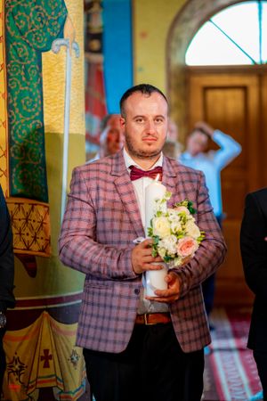 Groomsman in plaid blazer and bow tie holds white floral bouquet during church ceremony.