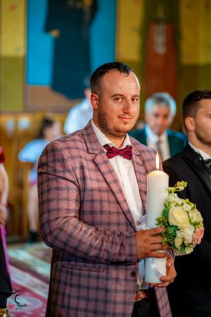 Groomsman in checkered pink suit and bow tie holds candle and flowers during ceremony.